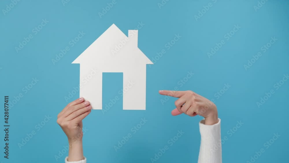 Close up of young female hand raising and pointing to small white paper house, posing isolated on blue studio background wall with copy space for advertisement. Property and mortgage concept Close up of young female hand raising and pointing to small white paper house, posing isolated on blue studio background wall with copy space for advertisement. Property and mortgage concept