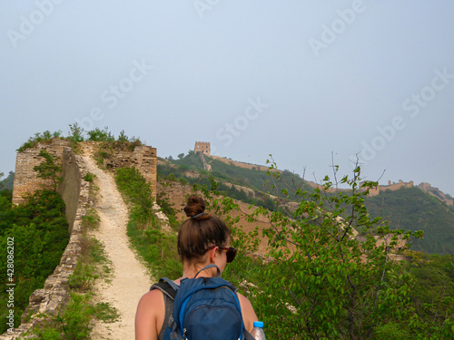 A woman with hiking backpack walking around a unrenewed Gubeikou part of Great Wall of China. The wall is spreading on tops of mountains. Dense forest around it. World wonder. Tradition and history photo