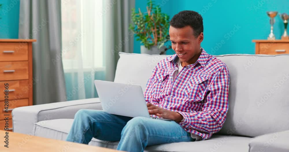 A handsome college student boy is studying remotely while sitting on couch in room at home. A teenager in a flannel plaid shirt is typing on laptop keyboard. A young man responds to an email A handsome college student boy is studying remotely while sitting on couch in room at home. A teenager in a flannel plaid shirt is typing on laptop keyboard. A young man responds to an email