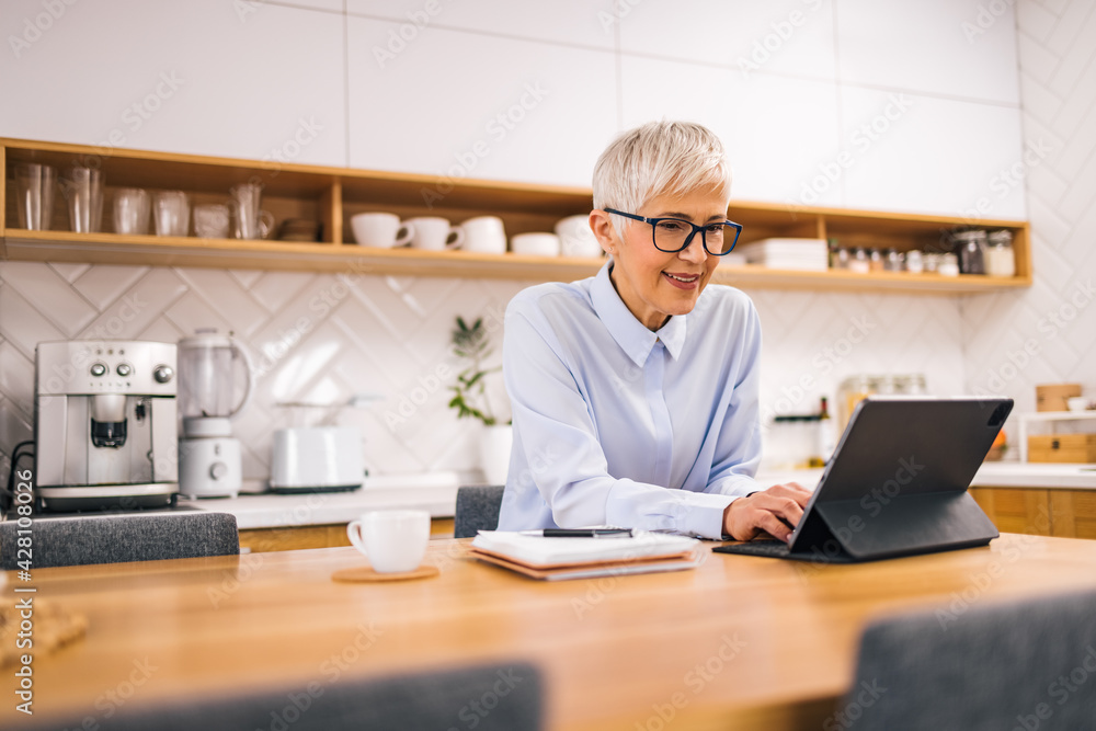 Portrait of a senior woman using tablet while sitting in the kitchen. Portrait of a senior woman using tablet while sitting in the kitchen.
