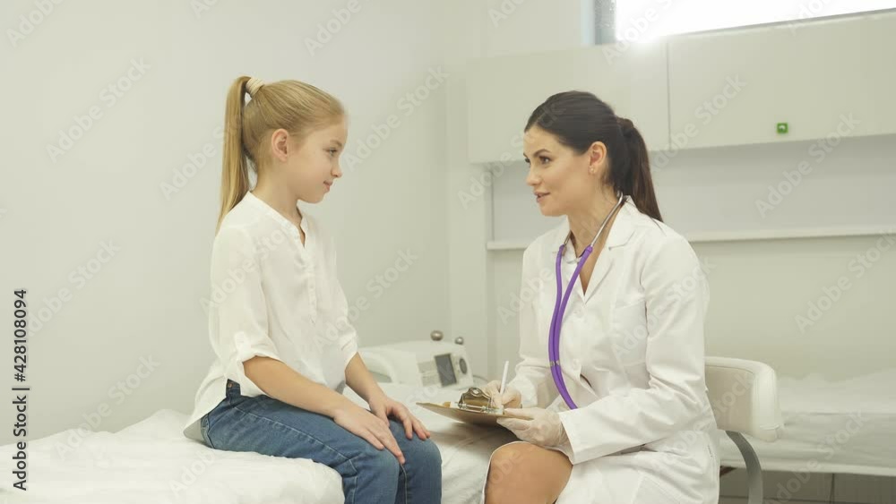 Female doctor pediatrician checking heartbeat of kid girl using stethoscope, in hospital room. close-up portrait of child looking up at doctor Female doctor pediatrician checking heartbeat of kid girl using stethoscope, in hospital room. close-up portrait of child looking up at doctor