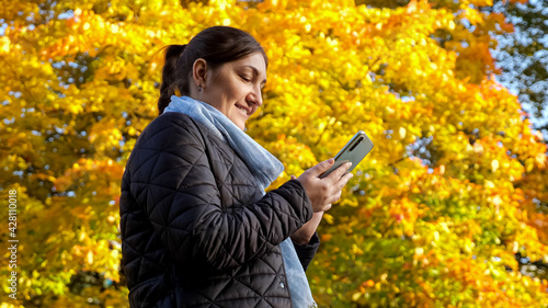 Young woman writes a message on the phone against the background of yellow trees.