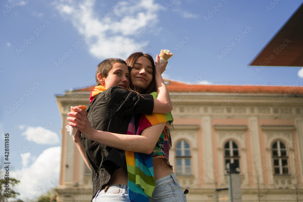 A young lesbian couple embraces their love. They are hugging on the street covered together with a rainbow colors scarf. A young lesbian couple embraces their love. They are hugging on the street covered together with a rainbow colors scarf.
