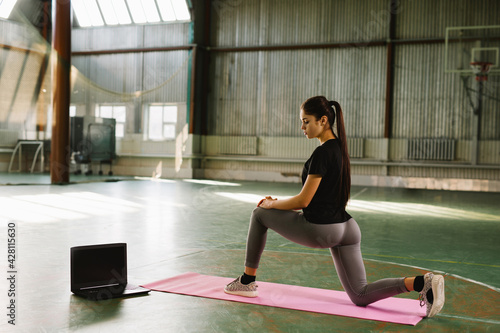 Sporty girl doing online yoga exercises on the mat in empty training gym during the quarantine period of coronavirus pandemic using laptop. An active healthy woman enjoys sports.