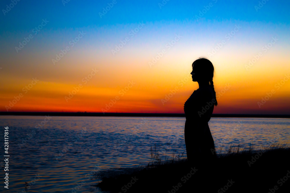 The girl at sunset stands at the shore and looks into the distance, waiting for a miracle.