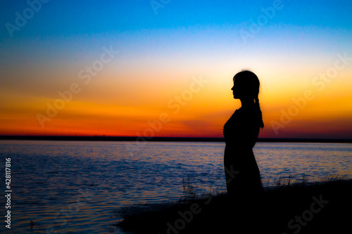 The girl at sunset stands at the shore and looks into the distance, waiting for a miracle.