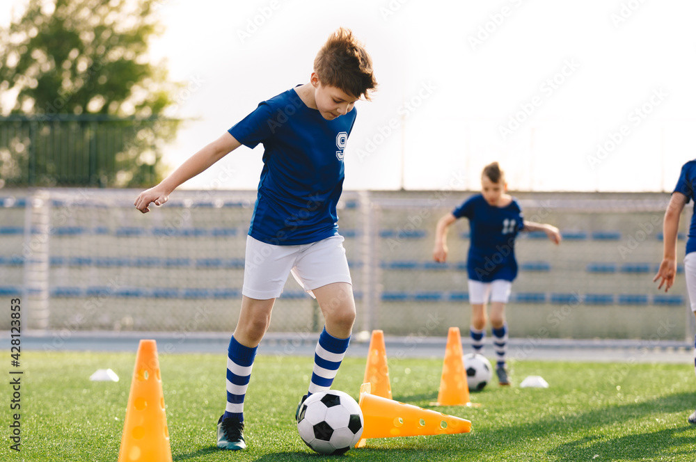 Obraz premium Happy young boy running and kicking the soccer ball in training. Group of school kids practicing football in the outdoor class. Youth soccer player knocking over the training cone.