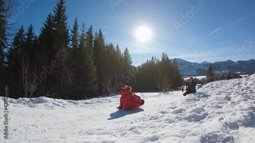 Kid in red jacket jump in slow motion on sled while sledding down from snowy hill in winter during sunny day in Zakopane.