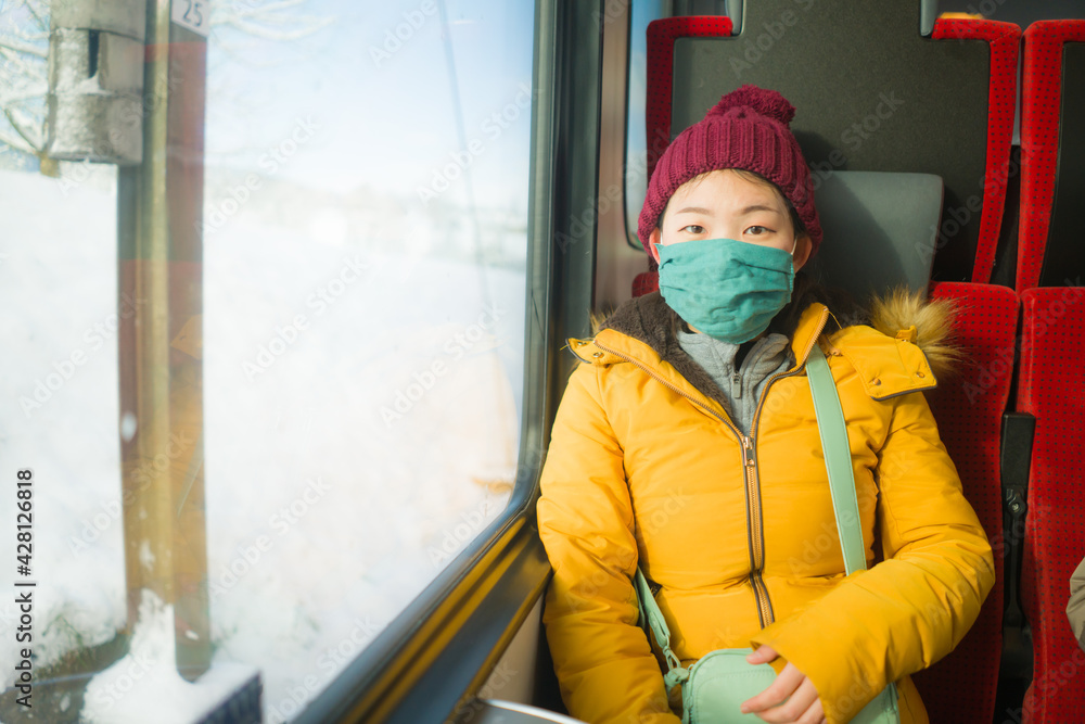 winter travel in times of covid19 - young happy and cute Asian Korean woman in face mask in the train enjoying snow landscape view at the window winter travel in times of covid19 - young happy and cute Asian Korean woman in face mask in the train enjoying snow landscape view at the window