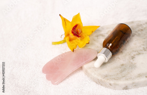 oil bottle or serum with gua sha stone at white towel background and yellow flowers. Beauty treatment concept. copy space