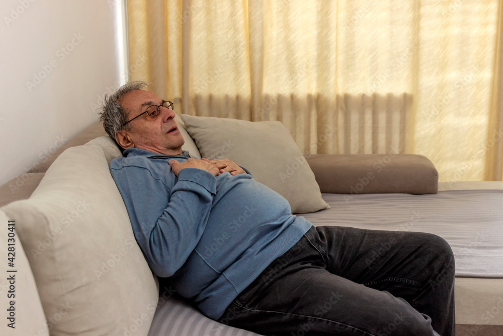 Shot of an uncomfortable looking old man holding his chest in discomfort while being seated on a couch at home. Overweight senior man holding breast because of heart infarction. Health care concept.
