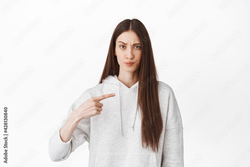Who me. Annoyed teenage girl frowning and pointing at herself, being accused, bothered as chosen to do something, staring displeased at camera, white background Who me. Annoyed teenage girl frowning and pointing at herself, being accused, bothered as chosen to do something, staring displeased at camera, white background