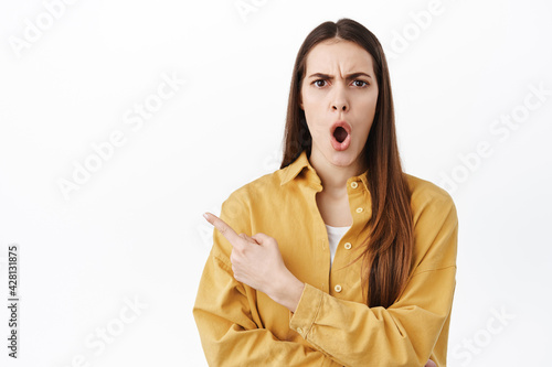 Shocked and frustrated woman complains, pointing finger aside at something offencive or rude, standing displeased and bothered against white background photo