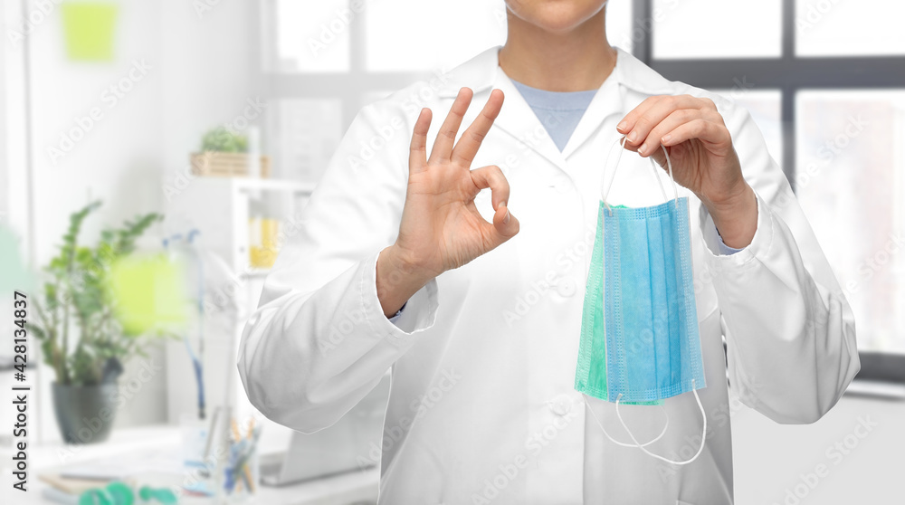 medicine, health protection and healthcare concept - close up of female doctor in white coat with medical mask showing ok gesture over medical office at hospital on background medicine, health protection and healthcare concept - close up of female doctor in white coat with medical mask showing ok gesture over medical office at hospital on background