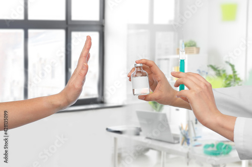 medicine, vaccination and healthcare concept - hand of doctor with syringe and drug in bottle and patient showing stop gesture over medical office at hospital on background photo