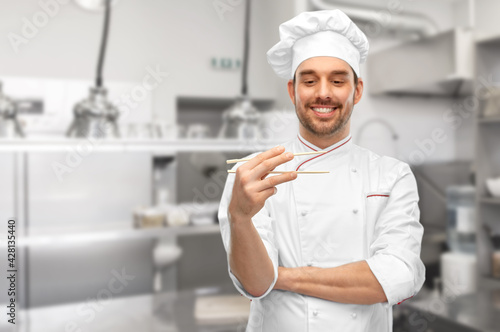 food cooking, asian cuisine and people concept - happy smiling male chef in toque holding something with chopsticks over restaurant kitchen background (focus on chopsticks)