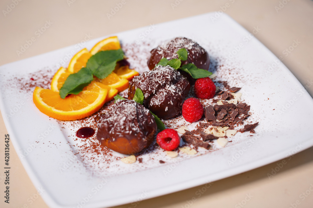 Three profiteroles covered with chocolate and coconut flakes decorated with orange and peanuts on a white plate Three profiteroles covered with chocolate and coconut flakes decorated with orange and peanuts on a white plate
