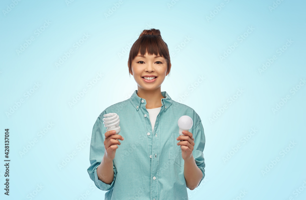 eco living, inspiration and sustainability concept - portrait of happy smiling young asian woman in turquoise shirt holding energy saving lighting bulb over blue background eco living, inspiration and sustainability concept - portrait of happy smiling young asian woman in turquoise shirt holding energy saving lighting bulb over blue background
