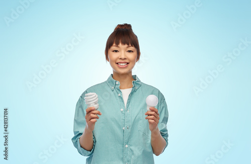 eco living, inspiration and sustainability concept - portrait of happy smiling young asian woman in turquoise shirt holding energy saving lighting bulb over blue background