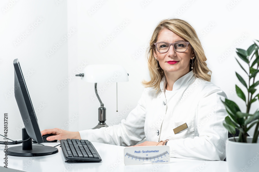 Blonde female physician doctor standing in Her office with Pedi-Scoliometer on a table. Portrait of friendly smiling woman-physician. Medicine concept Blonde female physician doctor standing in Her office with Pedi-Scoliometer on a table. Portrait of friendly smiling woman-physician. Medicine concept