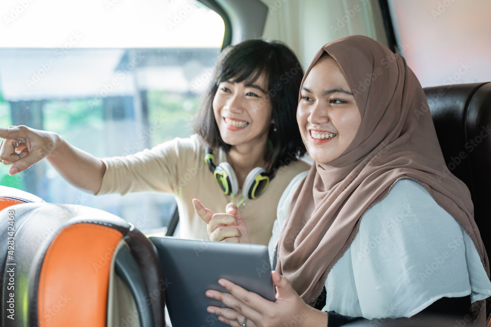 Fototapeta premium two young women smiling with pointing fingers while chatting while holding a tablet sitting on the bus