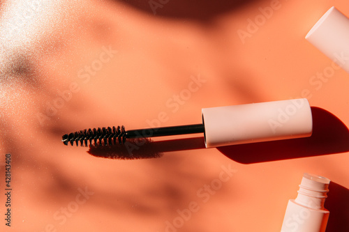 pink clean makeup brush for mascara lies next to an open tube, closed tubes of cosmetics, lip gloss, liquid lipstick, pink eyeliner on a peach background with shadows. Copy space