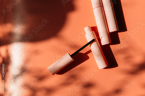 open tube with eyeliner brush next to closed pink tubes with mascara and liquid lipstick, lip gloss on a peach background with shadows
