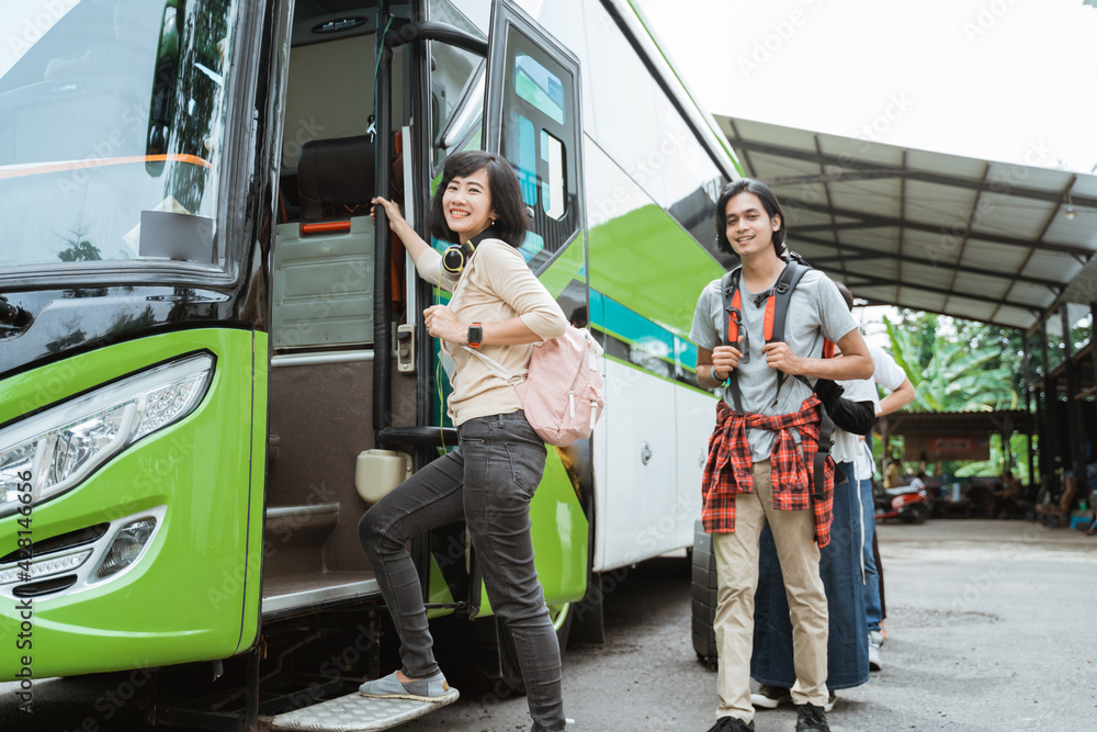 Asian woman carrying a backpack and headphones while holding the door handle gets into the bus with the background of passengers lining up to get on the bus