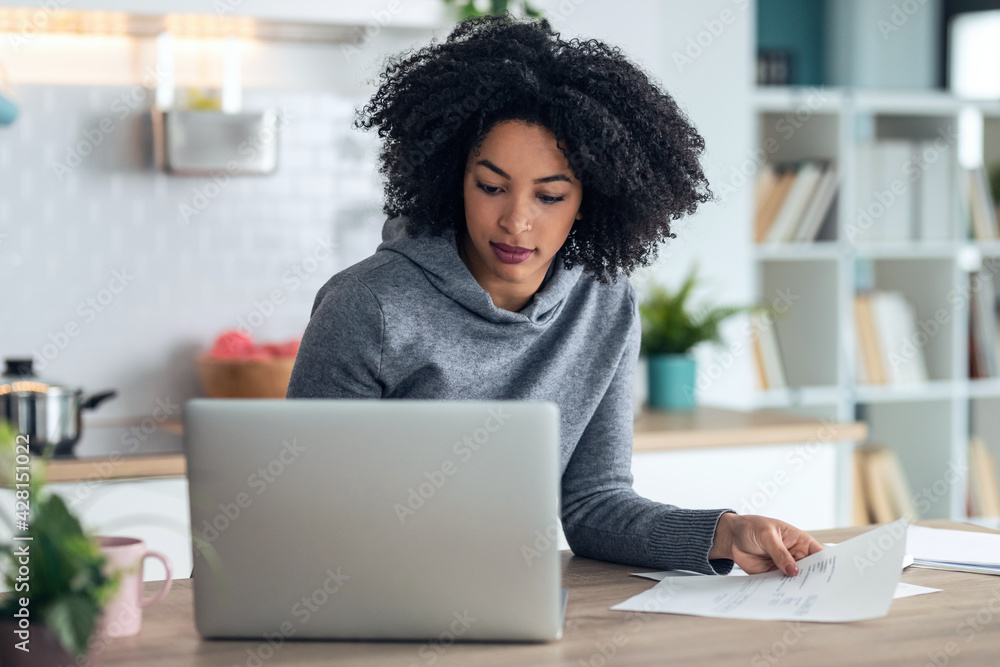 Afro business woman working with computer while consulting some invoices and documents in the kitchen at home. Afro business woman working with computer while consulting some invoices and documents in the kitchen at home.
