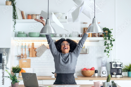 Fotografiet Happy afro business woman tossing invoices and documents into the air while working on the laptop at home