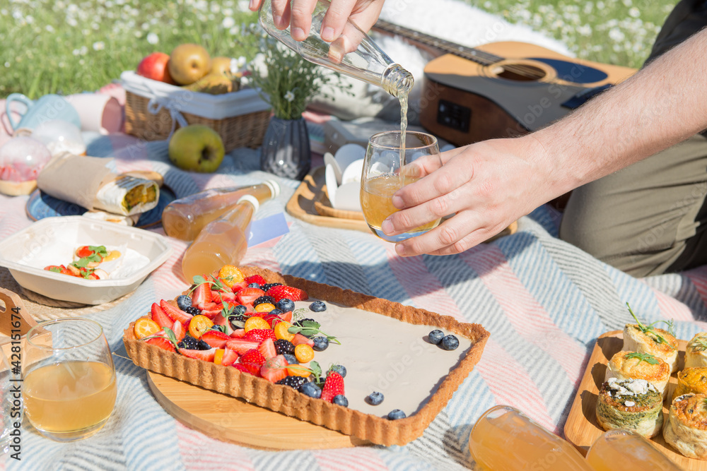 Naklejka premium Picnic setting with burgers, tart, croissant, cakes, picnic hamper basket, guitar and food ready for party. Cheerful family sitting on the grass during a picnic in a park. Young smiling family