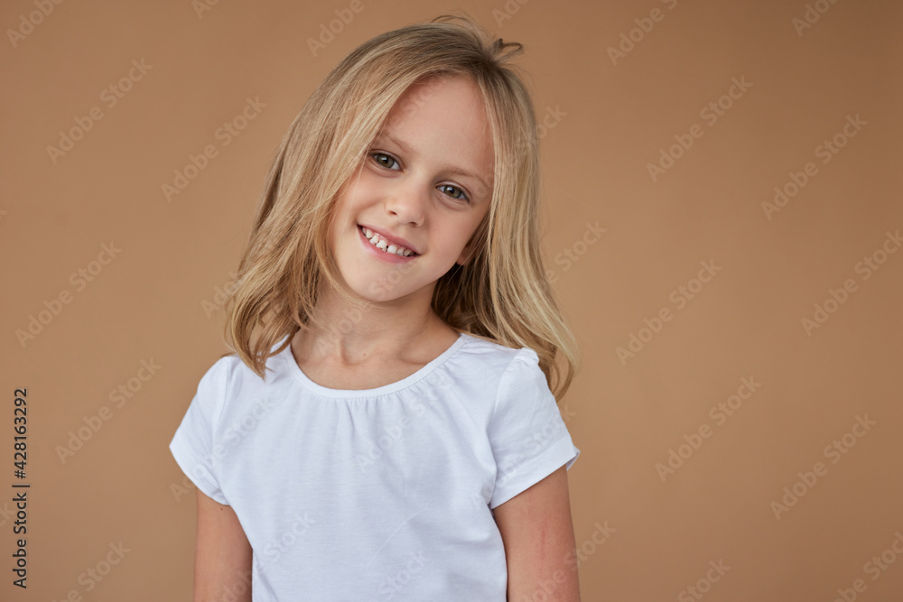 Close-up front portrait of pretty little girl with wavy blond hair, dressed in white clothes, over beige background. Close-up front portrait of pretty little girl with wavy blond hair, dressed in white clothes, over beige background.