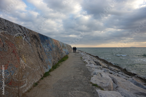 Paesaggio marino con figure umane : fidanzati che passeggiano in lontananza verso il centro dell'inquadratura , con mare a destra sotto gli scogli e a sinistra con murales colorati , cielo con nuvole photo