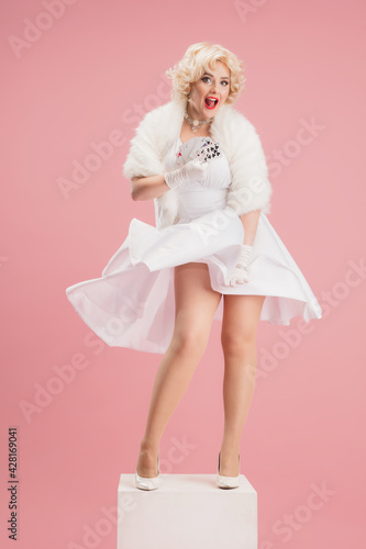 Portrait of young woman in white dress on coral pink background. Female model as a legendary actress. Pin up. Concept of comparison of eras, modern, fashion, beauty.