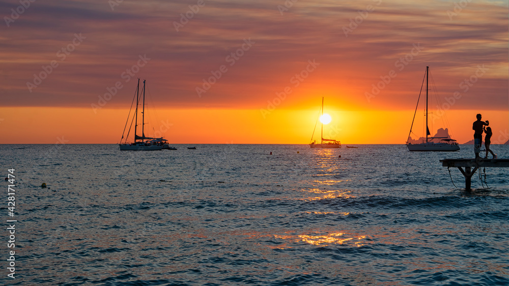 A couple relaxes watching the sunset over the sea from the pier and the silhouettes of the sailboats moored in the island of Formentera