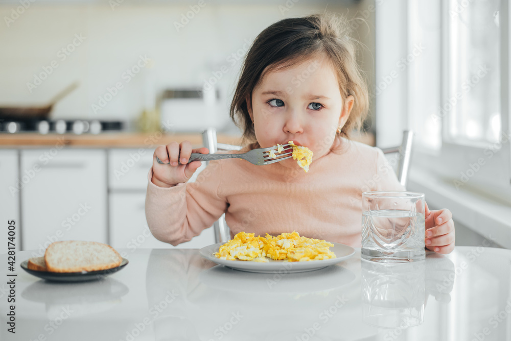 Baby girl in the afternoon in the white light kitchen in a pink sweater eating an omelet Baby girl in the afternoon in the white light kitchen in a pink sweater eating an omelet