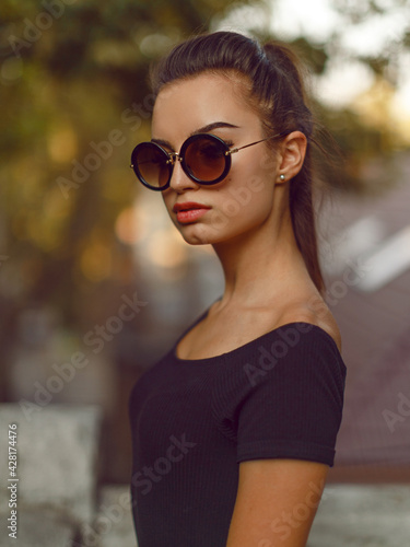 Frontal portrait of a beautiful brunette girl in black slim dress and sunglasses, hair arranged in ponytail.