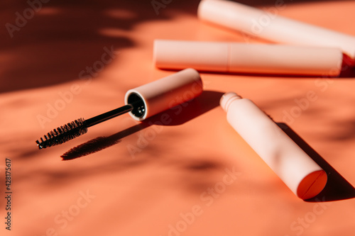 pink clean makeup brush for mascara lies next to an open tube, closed tubes of cosmetics, lip gloss, liquid lipstick, pink eyeliner on a peach background with shadows. Copy space