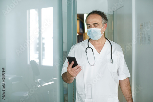 Medico cinquantenne in camice bianco e mascherina chirurgica guarda il suo cellulare  nella corsia di un ospedale  photo