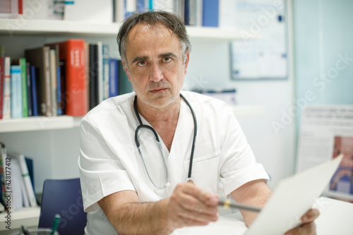 Medico cinquantenne in camice bianco seduto nella scrivania del suo ufficio guarda serio, mentre mostra un foglio.
