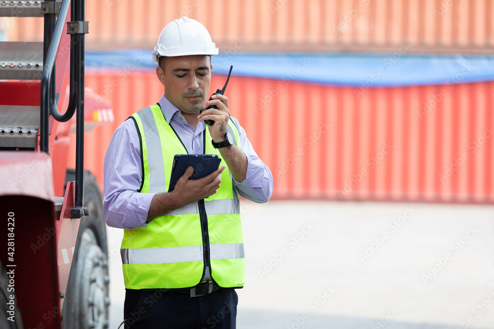 factory worker or engineer using walkie talkie and tablet for preparing a job beside truck in containers warehouse storage