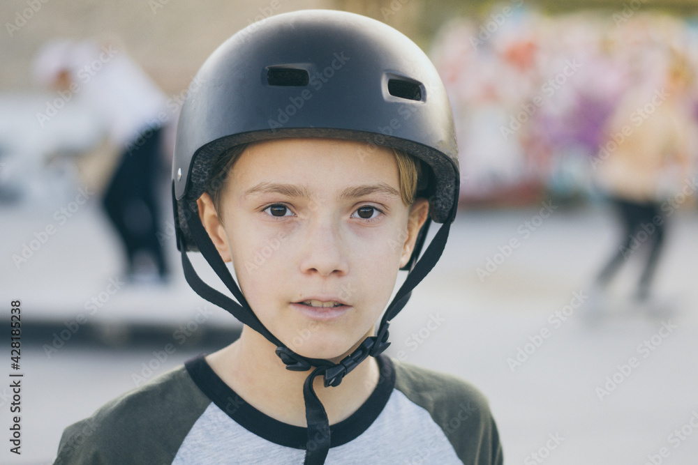 Portrait of trendy young skater at the skatepark wearing helmet, looking in camera. Teenager enjoying sunny day outdoors in the city with skateboard. Youth, happiness, safety, sport, positive concept Portrait of trendy young skater at the skatepark wearing helmet, looking in camera. Teenager enjoying sunny day outdoors in the city with skateboard. Youth, happiness, safety, sport, positive concept