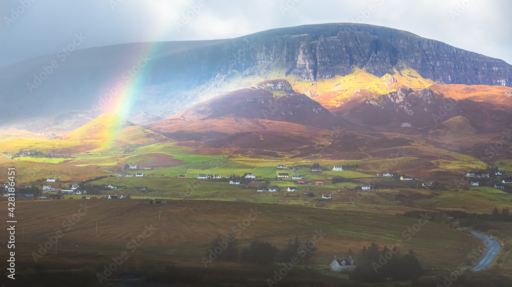 Moody, dramatic light and shadow with a colourful rainbow over the rural countryside landscape of the Quiraing at Trotternish ridge above Staffin village on the Isle of Skye, Scotland.