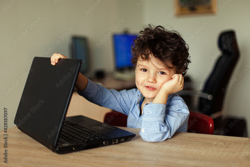 A cute curly boy sits at his laptop in the room. A preschool-age child is engaged in online training at home computer, distance learning, preparation for school