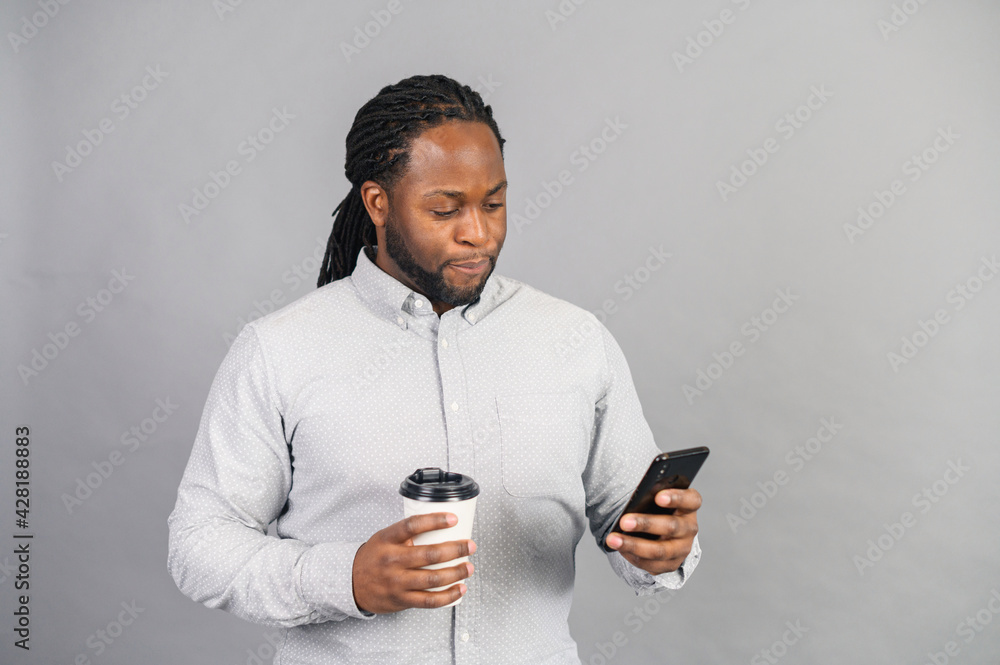 Serious, busy and confident African-American businessman wearing shirt and eyeglasses texting on the smartphone, holds a paper cup of take-away coffee, male office employee isolated Serious, busy and confident African-American businessman wearing shirt and eyeglasses texting on the smartphone, holds a paper cup of take-away coffee, male office employee isolated