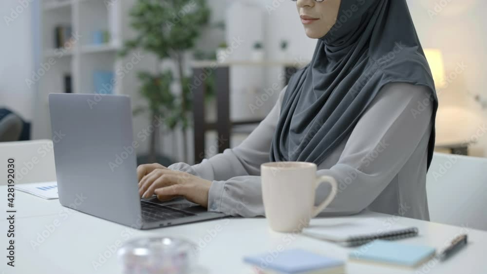 Concentrated muslim woman in headscarf and glasses working on laptop at home Concentrated muslim woman in headscarf and glasses working on laptop at home