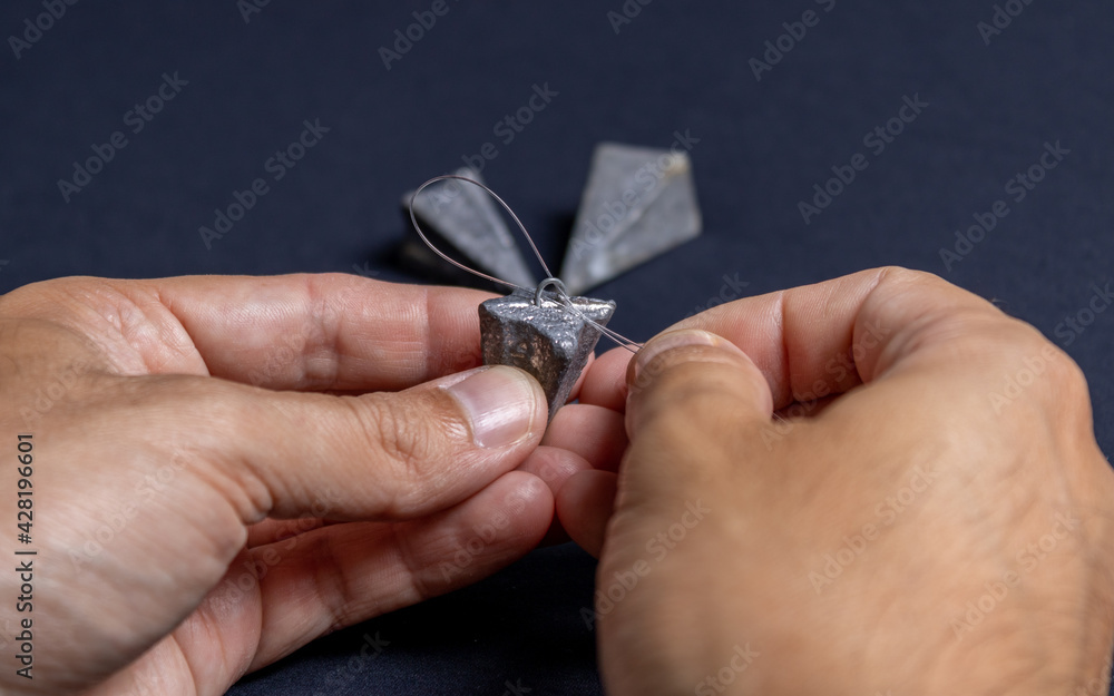 Fototapeta premium Fishing line and sinker being handled by the hand of a brown man demonstrating how to correctly thread the line on a black and dark background. Used for fishing and family leisure.