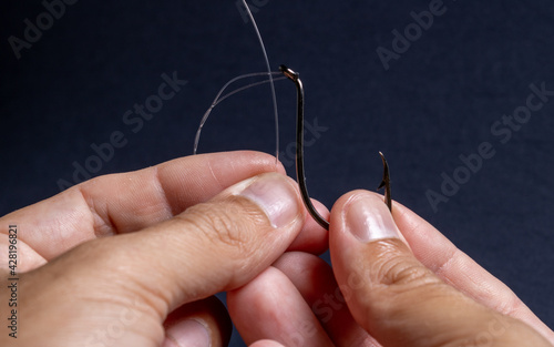 Fishing line and hook being handled by the hands of a brown man demonstrating how to thread the line correctly on a black and dark background. Used for fishing and family leisure.