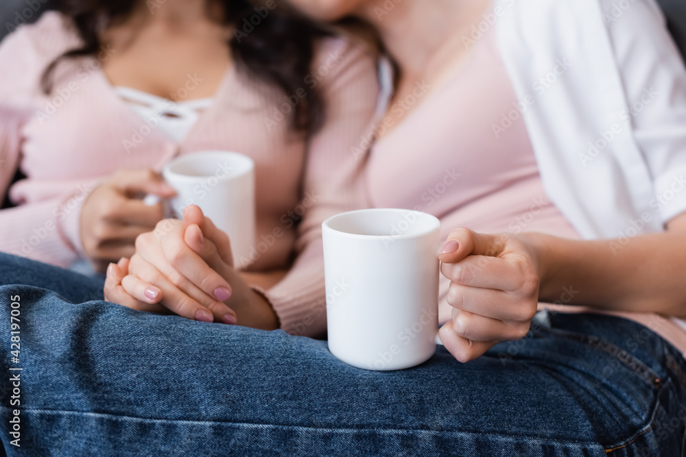cropped view of girlfriends holding cups of tea in living room cropped view of girlfriends holding cups of tea in living room