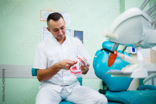 Positive doctor in white uniform showing mock jaw with teeth while sitting in dental office